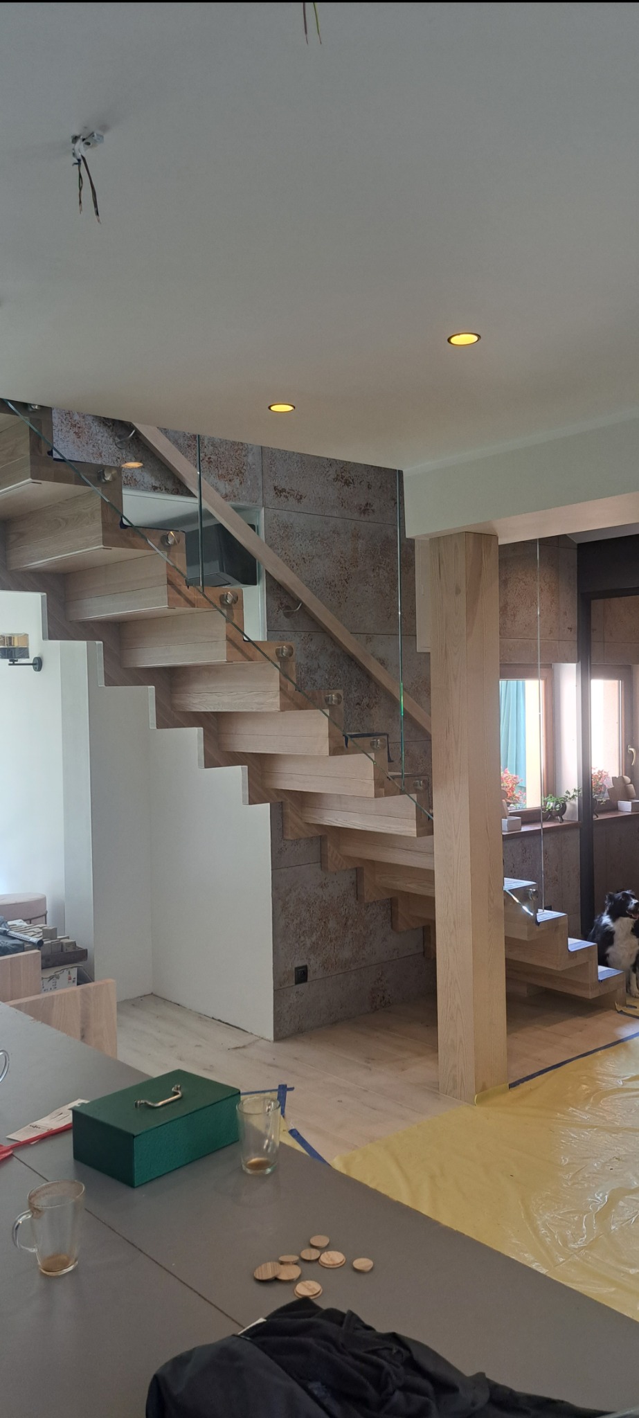 Nowoczesne schody z jasnego drewna i szklanymi balustradami w jasnym wnętrzu. Widoczny fragment blatu z narzędziami i pies.