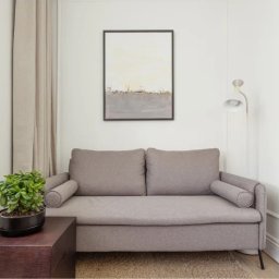 Holy Moly Terapie Anna Bielańska - Jasnoszara sofa z poduszkami w minimalistycznym wnętrzu. Obraz w ramie na ścianie, roślina w doniczce na stoliku, lampa podłogowa. Stonowana kolorystyka.