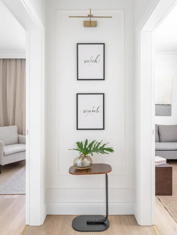 Jasne wnętrze: minimalistyczny hol z dekoracyjnymi grafikami, subtelnym oświetleniem i rośliną w szklanym wazonie na stoliku. Widok na pokoje po obu stronach.