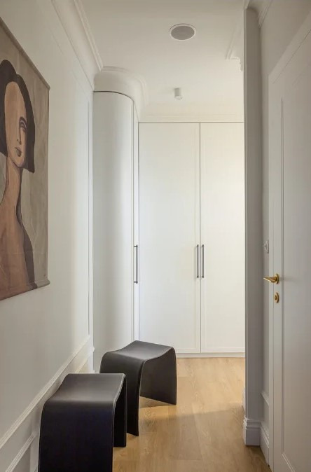 Minimalistyczny przedpokój z dwoma czarnymi taboretami, obrazem na ścianie i białymi szafami w zabudowie. Jasne drewno na podłodze. Nowoczesne i eleganckie wnętrze.