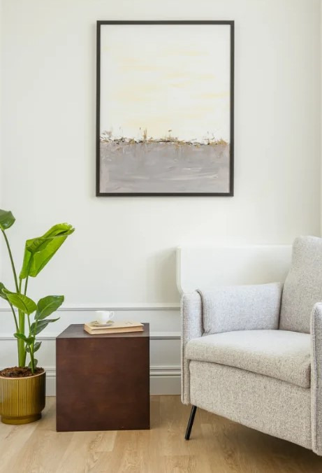 Jasny pokój z abstrakcyjnym obrazem w czarnej ramie nad fotelem. Na stoliku obok stoi roślina doniczkowa i filiżanka na książce. Minimalistyczny wystrój wnętrza.