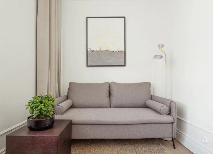 Jasnoszara sofa z poduszkami w minimalistycznym wnętrzu. Obraz w ramie na ścianie, roślina w doniczce na stoliku, lampa podłogowa. Stonowana kolorystyka.