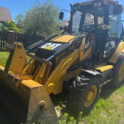 Żółta koparko-ładowarka CAT 432F na trawie, w tle ogrodzenie i dom. Sprzęt budowlany gotowy do pracy.