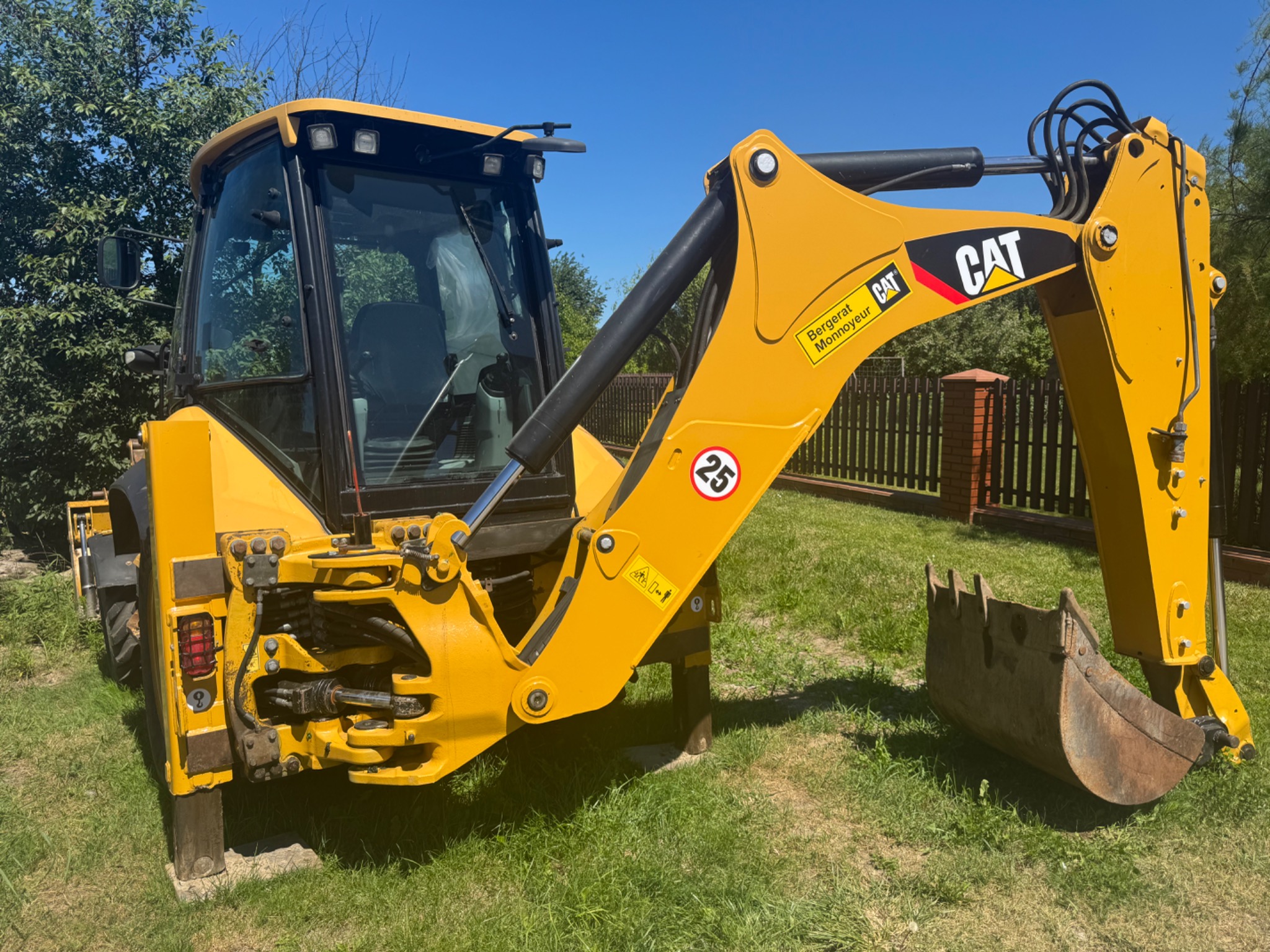 Żółta koparko-ładowarka CAT na trawie, z widocznym ramieniem kopiącym i kabiną operatora, na tle ogrodzenia i drzew, w słoneczny dzień.