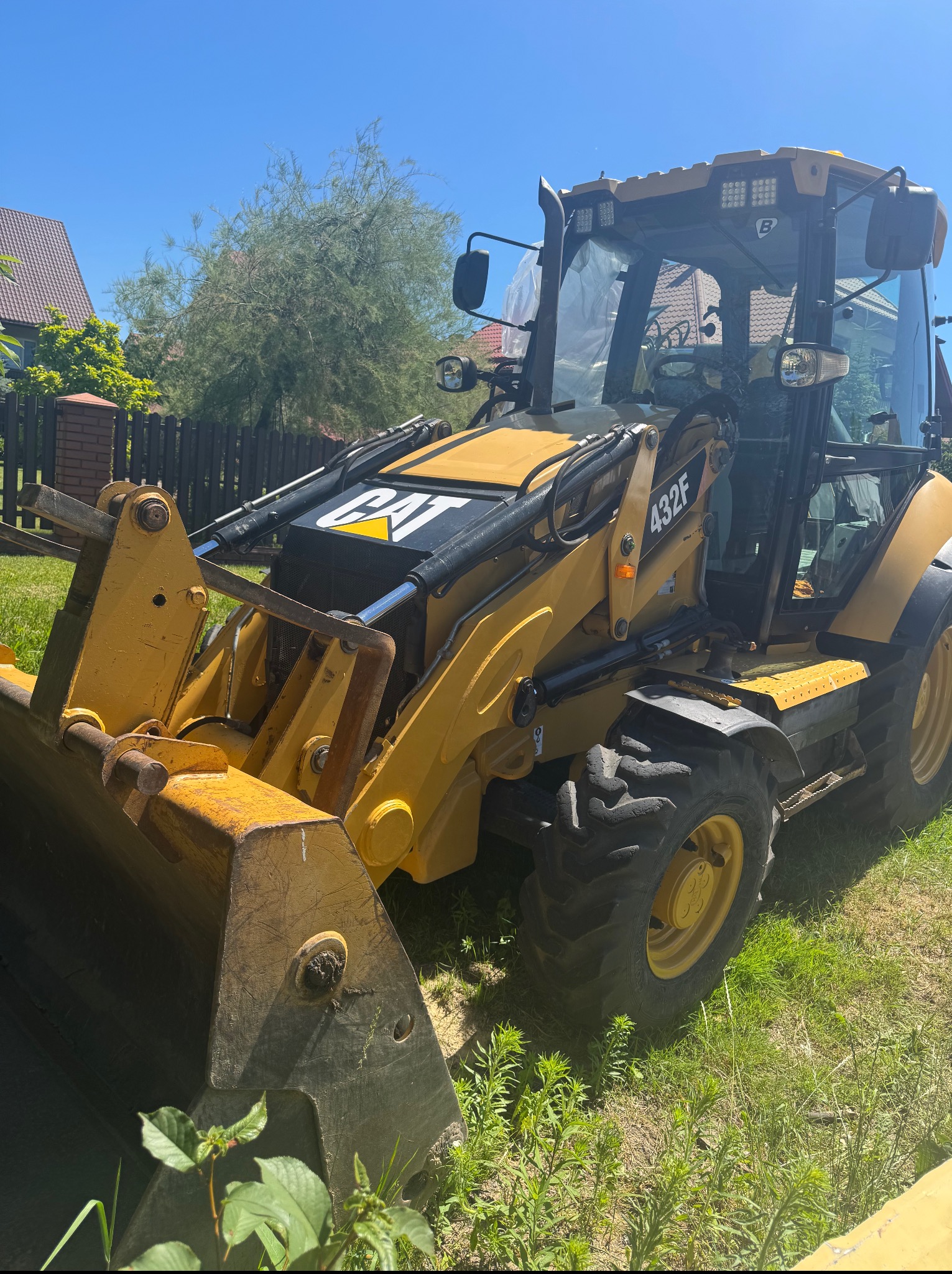 Żółta koparko-ładowarka CAT 432F na trawie, w tle ogrodzenie i dom. Sprzęt budowlany gotowy do pracy.