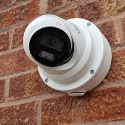 Biała kamera monitoringu kopułkowa zamontowana na ceglanej ścianie zewnętrznej. Widoczne śruby mocujące i oznaczenie kierunku 'UP'.