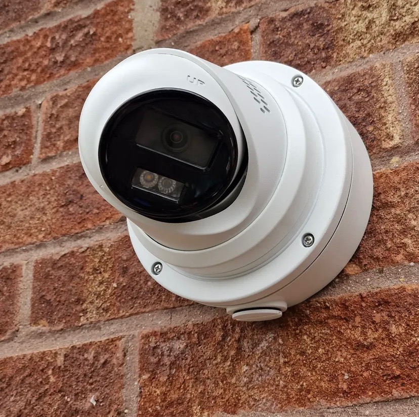 Biała kamera monitoringu kopułkowa zamontowana na ceglanej ścianie zewnętrznej. Widoczne śruby mocujące i oznaczenie kierunku 'UP'.