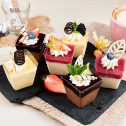 CENTRAL FUND OF IMMOVABLES SPÓŁKA Z OGRANICZONĄ ODPOWIEDZIALNOŚCIĄ - Deserowe mini-porcje na czarnym łupku: kremy, galaretki, owoce i czekoladowe dekoracje w przezroczystych kostkach. Obok filiżanka cappuccino i srebrna łyżeczka.