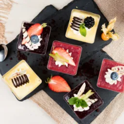Deserki w kwadratowych pojemnikach, udekorowane owocami i czekoladą, ułożone na czarnej łupkowej płycie. Obok filiżanka kawy i łyżeczka.