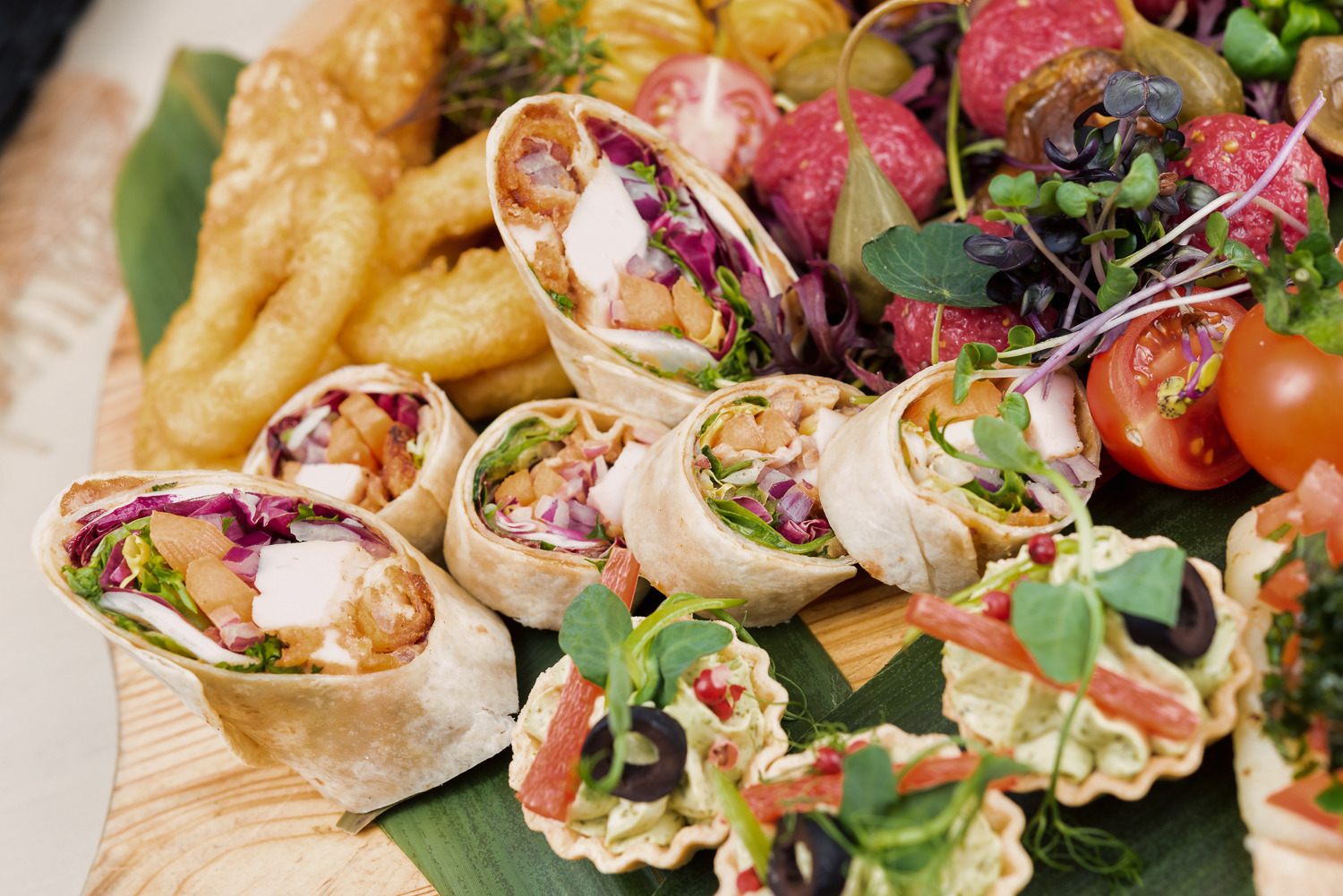 Dekoracyjna drewniana deska z roladkami tortilli z kurczakiem, mini tartaletkami i przekąskami, idealna na catering firmowy lub imprezę okolicznościową.