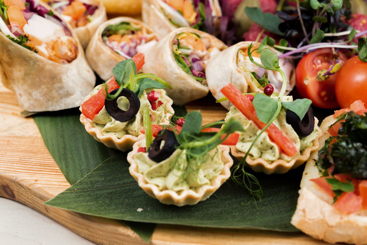 Dekoracyjne tartaletki z kremem i warzywami, obok tortille i bruschetta na drewnianej desce z liściem. Elegancki finger food na przyjęcie.