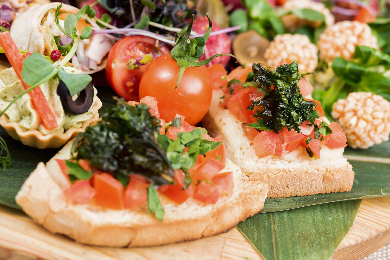Dekoracyjne przekąski na drewnianej desce z liściem bambusa: tartaletka z kremem, bruschetta z pomidorami i ziołami, mini tortille i kulki ryżowe.