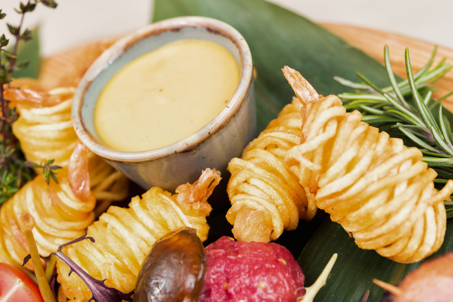 Kreatywna kompozycja kulinarna: krewetki w ziemniaczanych spiralach obok miseczki z sosem, całość na liściu z gałązką rozmarynu, z bliska.