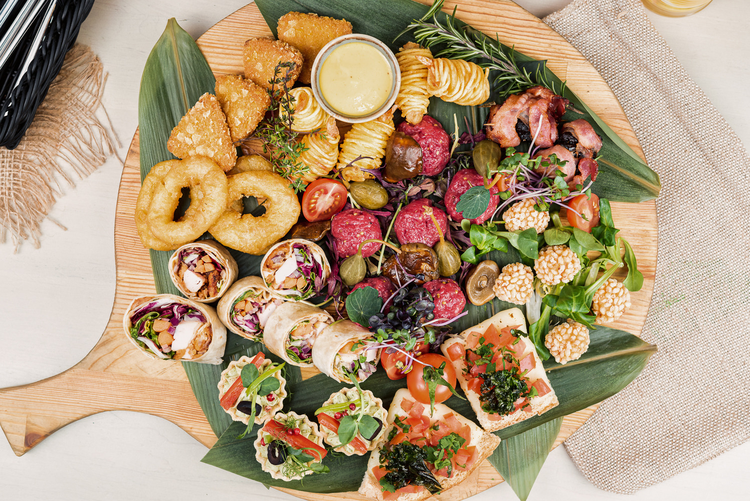 Dekoracyjna deska z przekąskami: tartinki, roladki, mini tarty, smażone krążki i mięsne kulki, ułożone na liściu i drewnianej desce. Różnorodność smaków i kolorów.