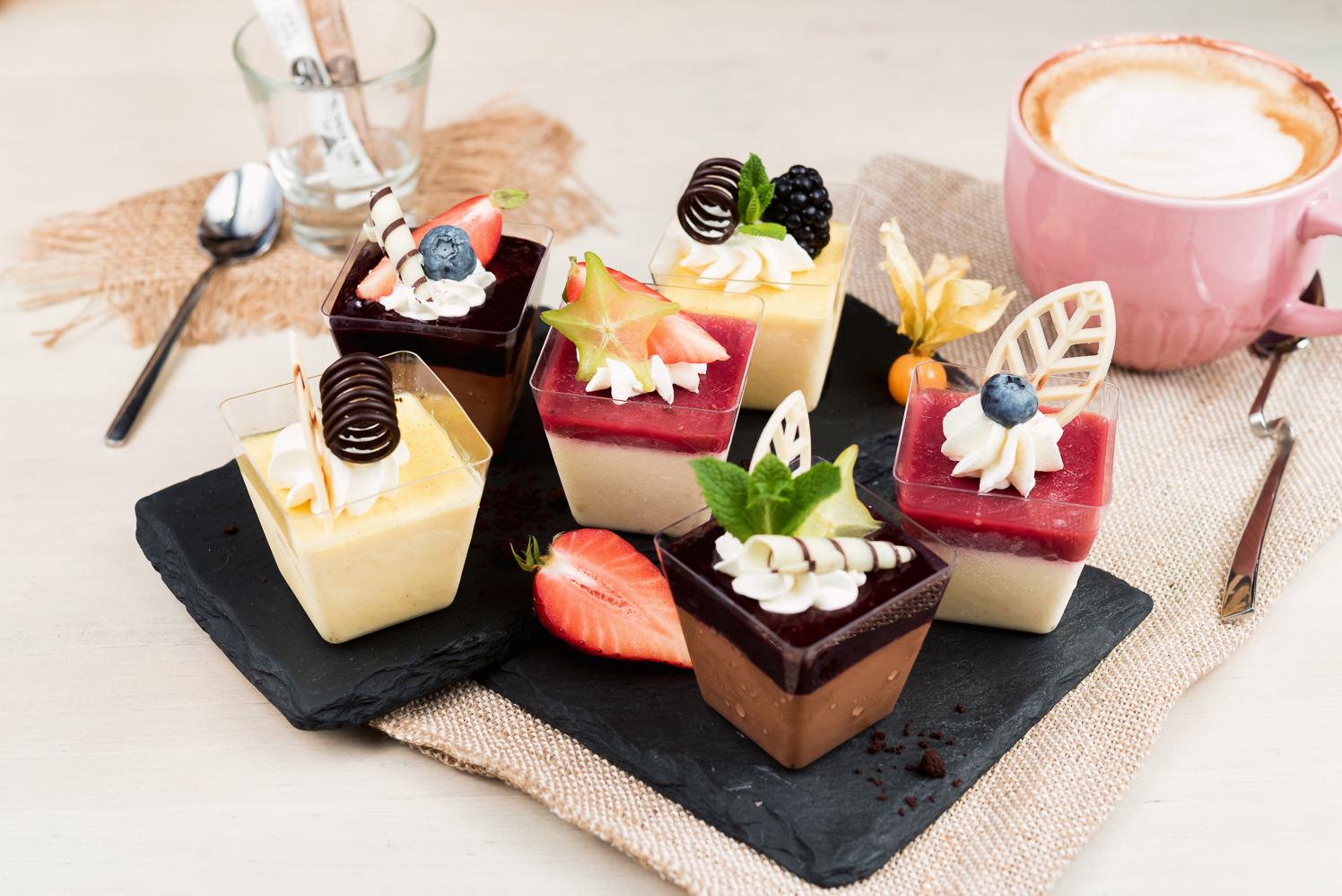 Deserowe mini-porcje na czarnym łupku: kremy, galaretki, owoce i czekoladowe dekoracje w przezroczystych kostkach. Obok filiżanka cappuccino i srebrna łyżeczka.