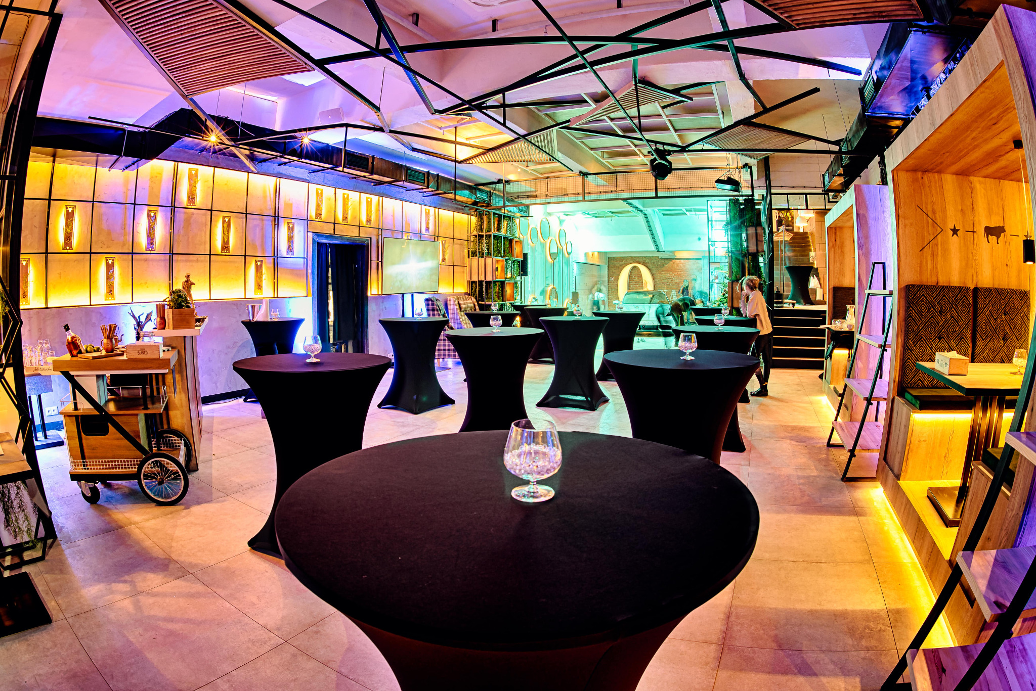 Elegancka sala bankietowa z czarnymi stolikami koktajlowymi i szklankami, oświetlona dekoracyjnym światłem. Widoczny bar na kółkach i osoba w tle.