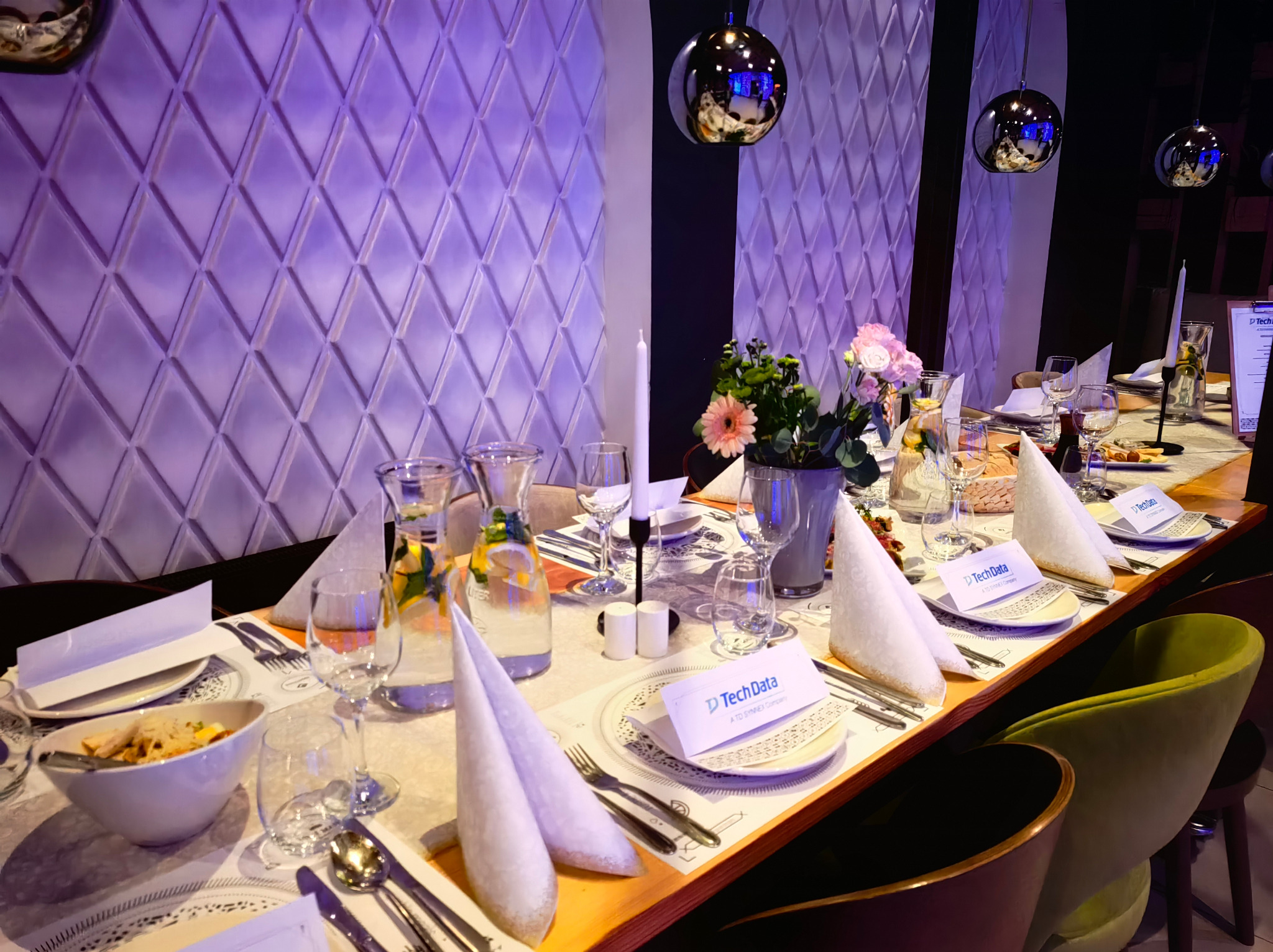 Elegancko nakryty stół z dekoracjami kwiatowymi, serwetkami w kształcie stożków i naczyniami, oświetlony dekoracyjnymi lampami wiszącymi, tworzący atmosferę wykwintnego przyjęcia.