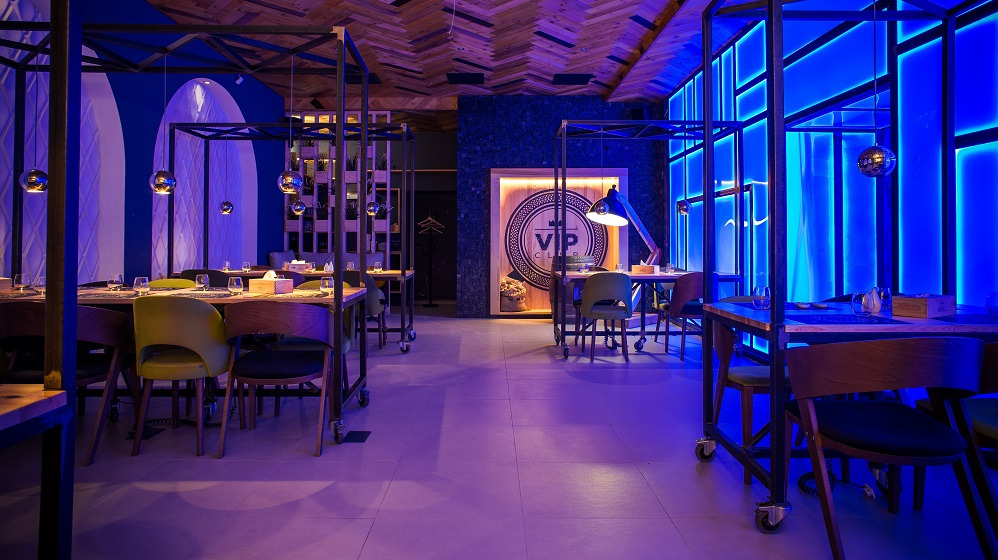 Elegancka sala restauracyjna z drewnianym sufitem i stołami nakrytymi dla gości, oświetlona niebieskim światłem. Widoczne dekoracje i logo VIP Club na ścianie.