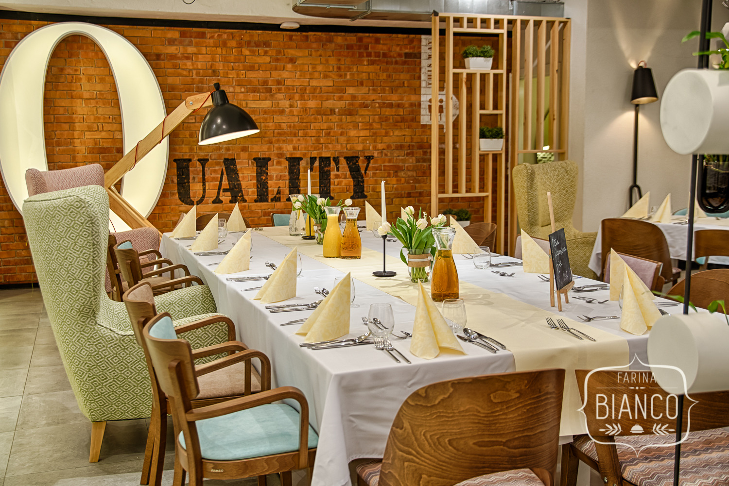 Uroczysty stół z beżowymi serwetkami, kwiatami i szklanymi naczyniami na tle ceglanej ściany z napisem 'Quality', oświetlony lampą.