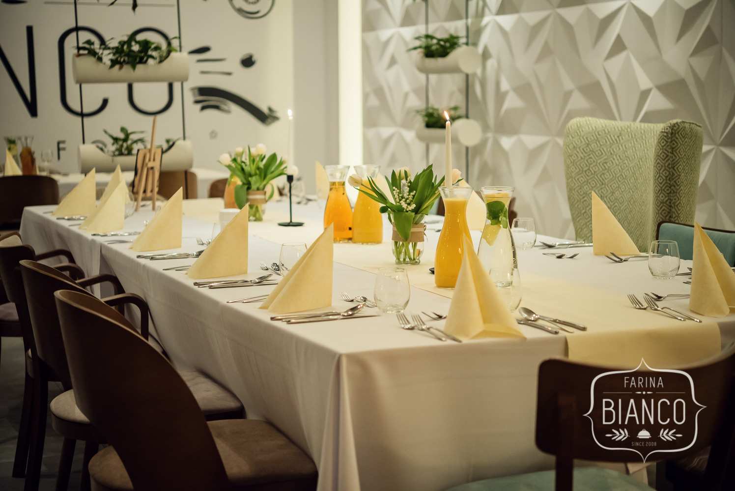 Elegancko nakryty stół z jasnymi serwetkami, kwiatami w wazonach i karafkami z napojami. W tle dekoracje ścienne i krzesła z logo Farina Bianco.