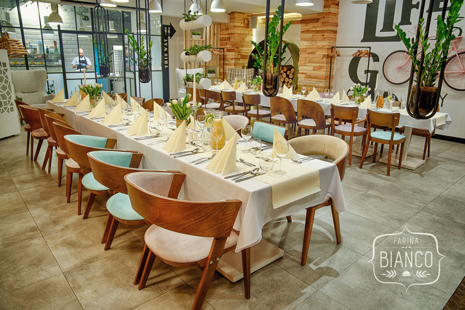 Uroczysta aranżacja stołu z kremowymi serwetami i elegancką zastawą w jasnym wnętrzu restauracji Farina Bianco, z widoczną kuchnią w tle.