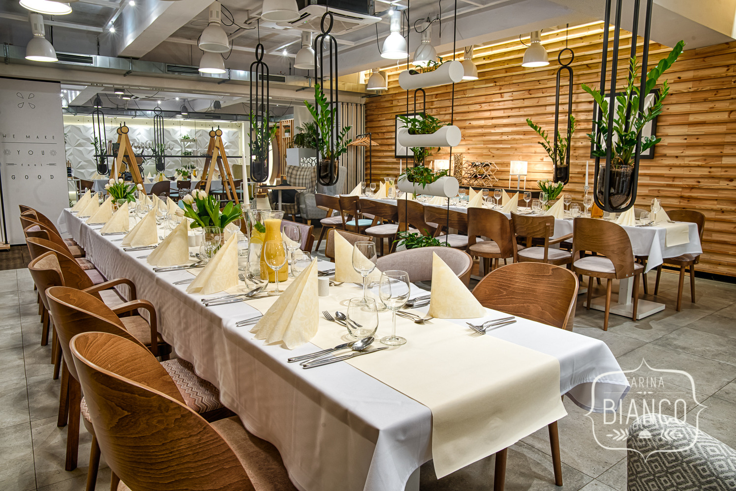 Elegancko nakryty stół z białym obrusem, beżowymi serwetkami i kwiatami w restauracji, drewniane krzesła i dekoracje roślinne w tle.