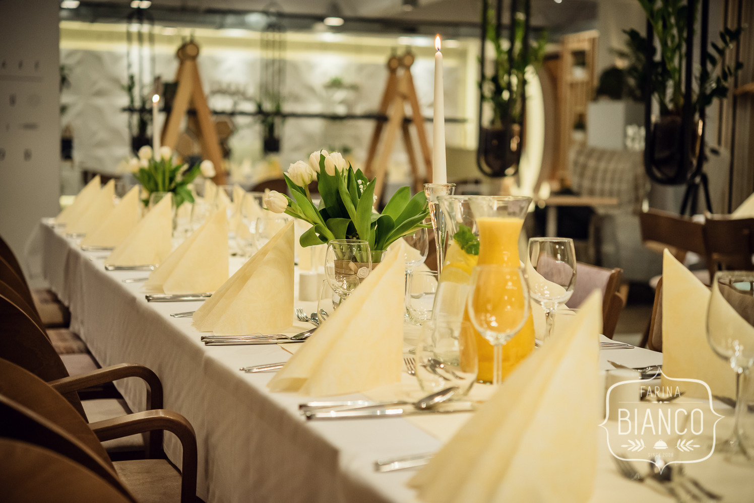 Elegancko nakryty stół z białym obrusem, kwiatami w wazonie, świecą i dzbankiem soku. W tle jasne wnętrze restauracji.