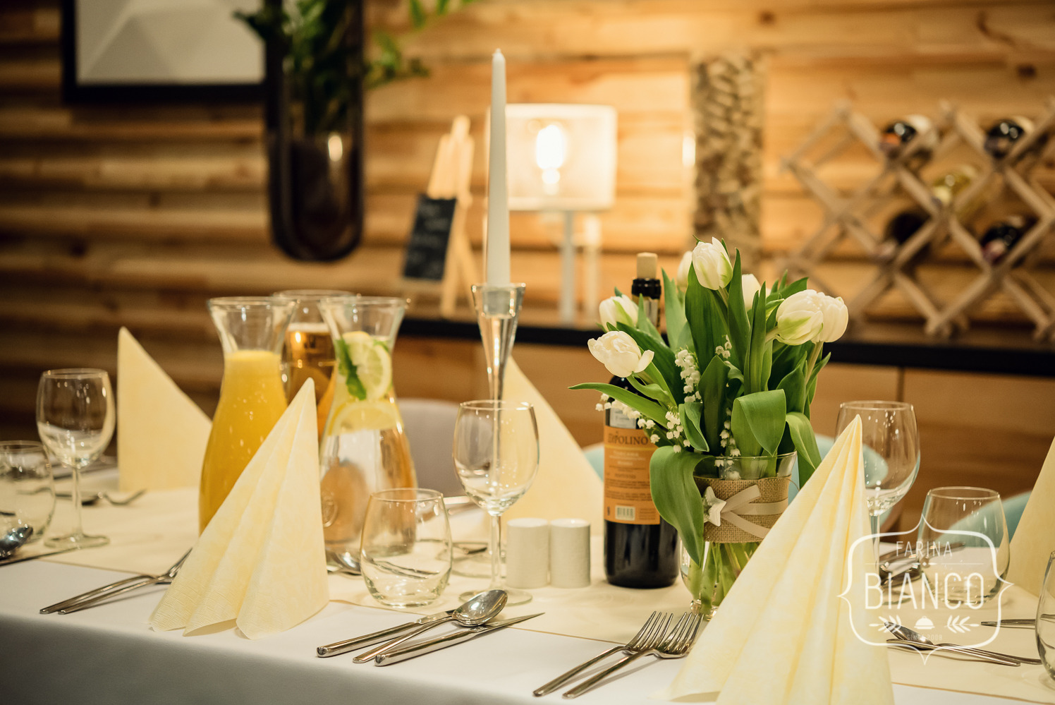 Elegancko nakryty stół z kwiatami, napojami i zastawą, gotowy na przyjęcie w rustykalnym stylu. W tle drewniana ściana i dekoracje.