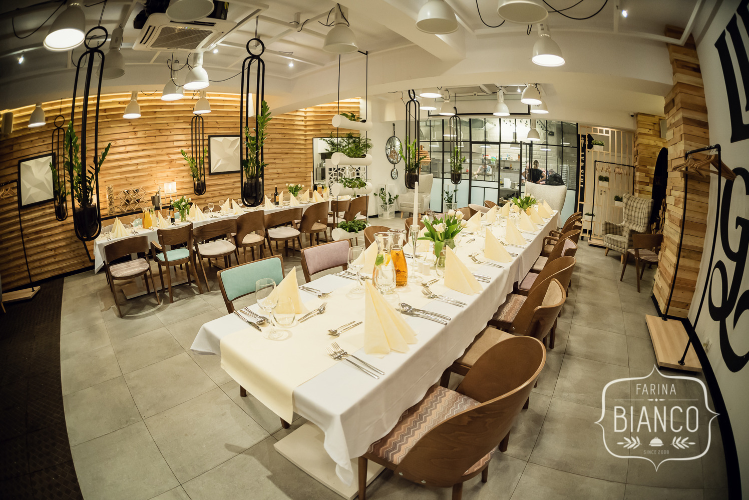 Elegancko nakryty stół w restauracji Farina Bianco, z białymi obrusami, serwetkami i tulipanami. W tle drewniane ściany i wiszące rośliny.