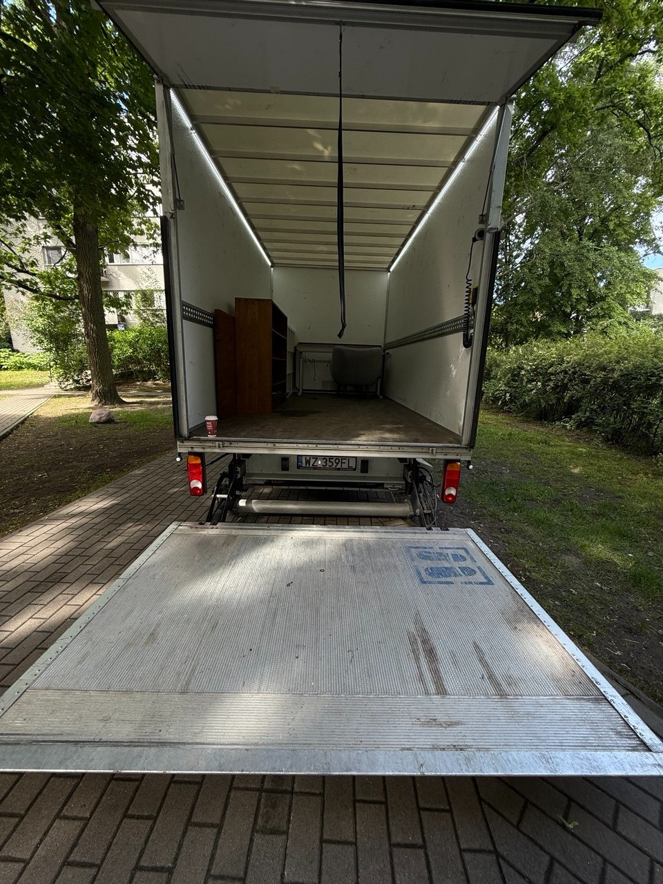 Otwarta paka busa z podniesioną rampą, widoczne meble wewnątrz, tablica rejestracyjna i otoczenie zieleni. Gotowość do załadunku podczas przeprowadzki.