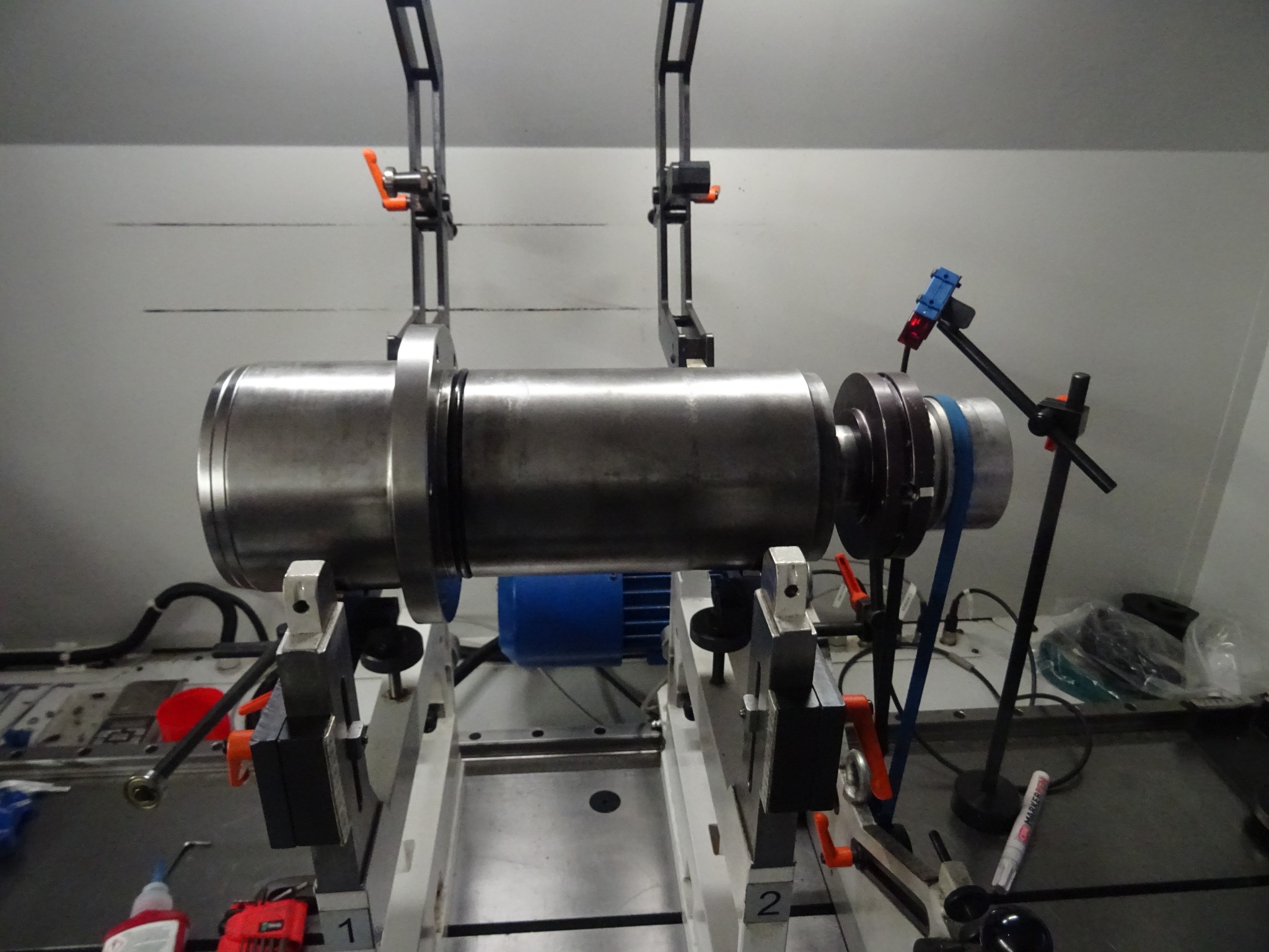 Stalowy wał wrzeciona w procesie regeneracji, zamontowany na specjalistycznym stanowisku pomiarowym z czujnikami i narzędziami. Widoczny silnik, przewody i akcesoria.