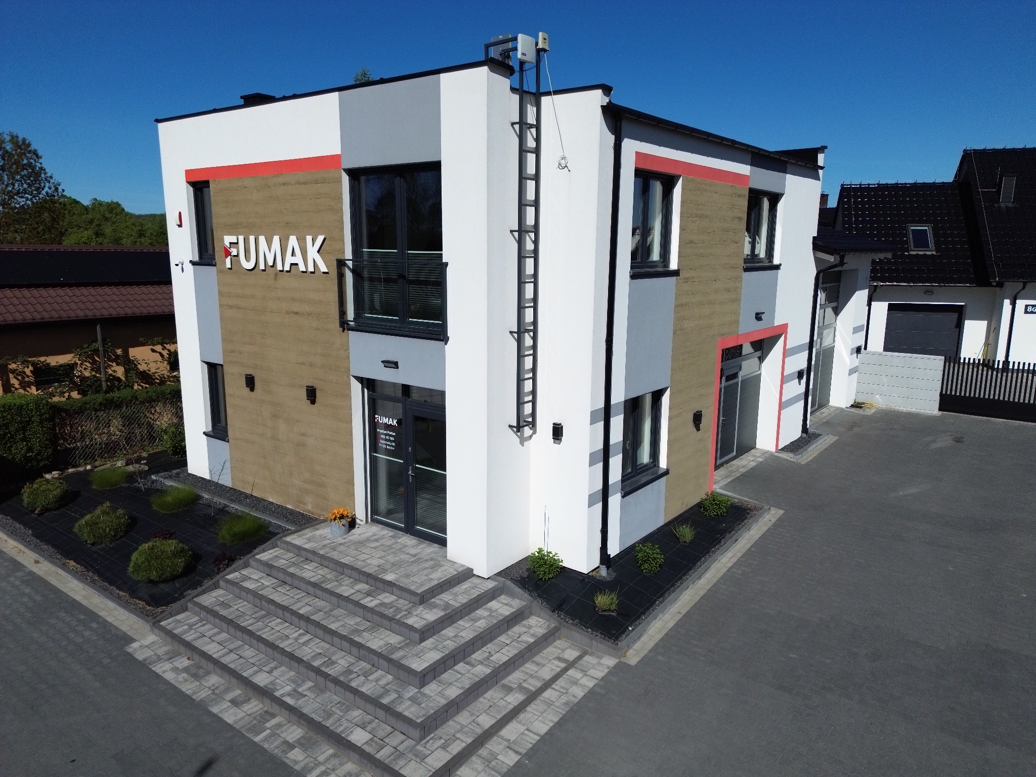 Nowoczesny budynek firmy FUMAK z jasną elewacją, schodami wejściowymi i elementami szarości, czerwieni. Na dachu drabina i instalacja. Widok z góry na kostkę brukową.