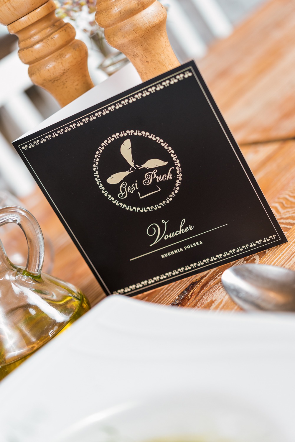 Elegancki voucher restauracji Gęsi Puch kuchni polskiej na drewnianym stole, obok oliwa i sztućce. Prezentacja oferty restauracji.