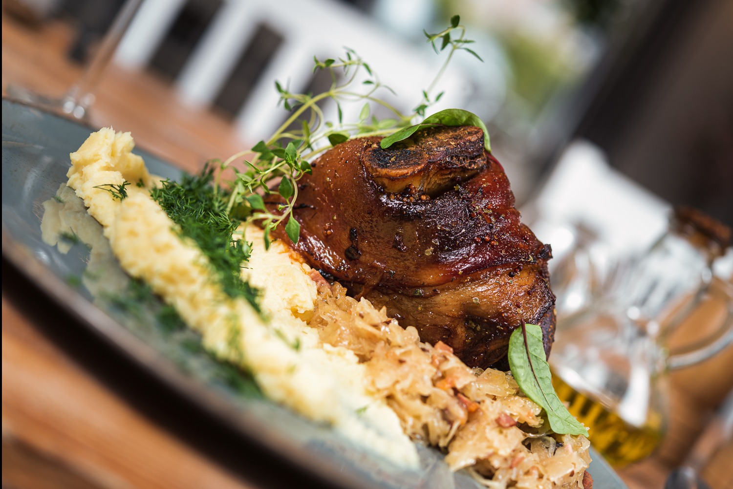 Pieczona golonka wieprzowa, podana na talerzu z puree ziemniaczanym i kapustą, udekorowana ziołami. Kąt ujęcia podkreśla apetyczny wygląd dania.