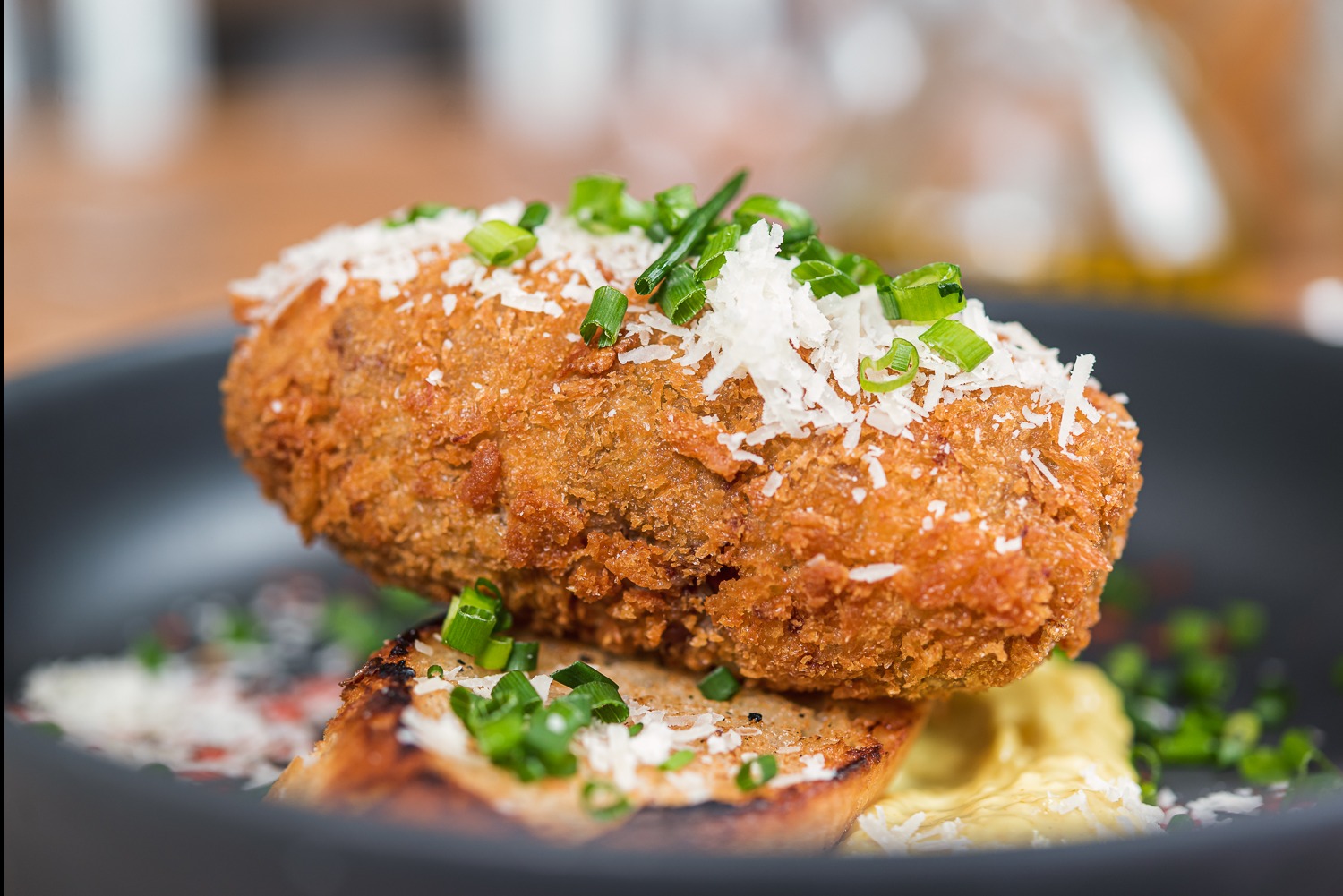 Krokiet z cielęciny na grzance, posypany serem i szczypiorkiem, podany na ciemnym talerzu z sosem. Elegancka aranżacja dania na przyjęcie.