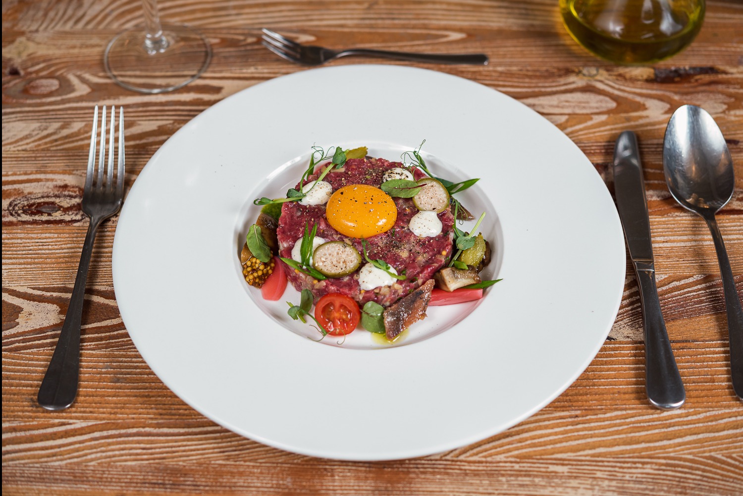Elegancko podany tatar wołowy z żółtkiem, warzywami i ziołami na białym talerzu, ustawiony na drewnianym stole z widelcem, nożem i łyżką. Fine dining.