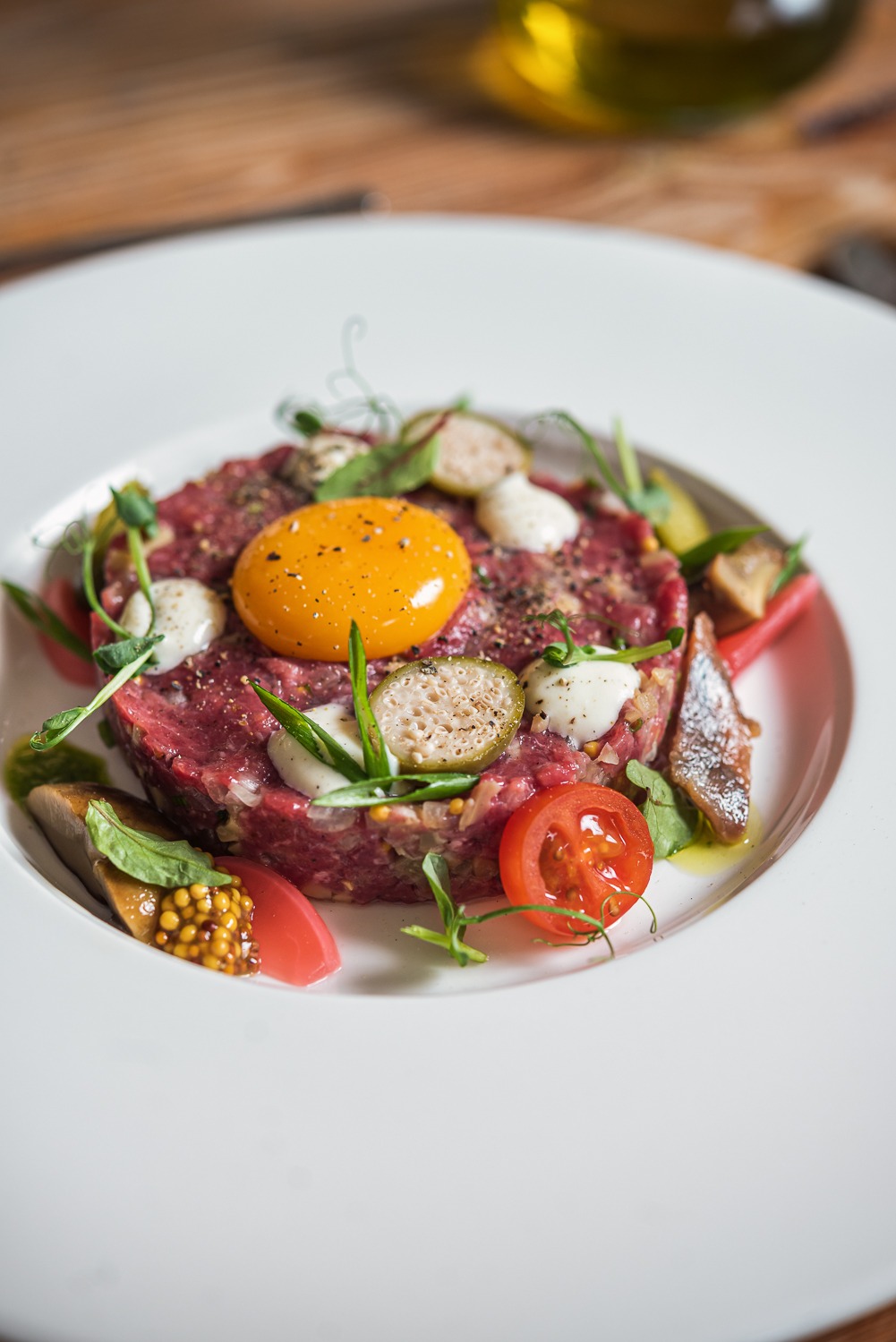 Elegancko podany tatar wołowy z żółtkiem, kaparami, marynowanymi grzybami i pomidorem na białym talerzu. Fine dining, zbliżenie.