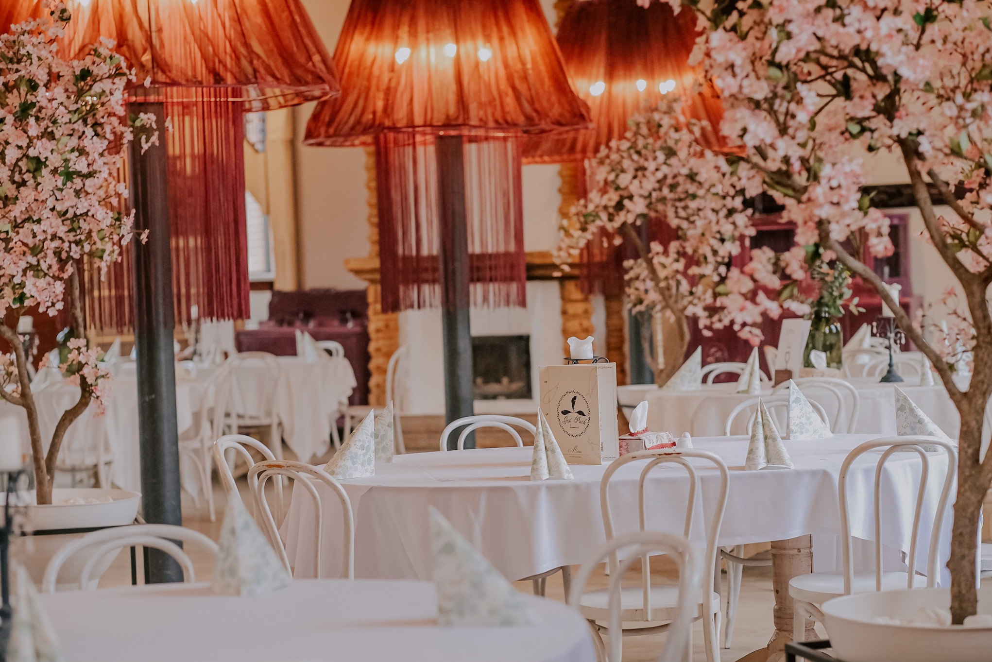 Sala bankietowa z białymi stołami, dekoracjami z kwiatów wiśni i lampami z frędzlami, przygotowana na przyjęcie. Elegancki wystrój wnętrza.