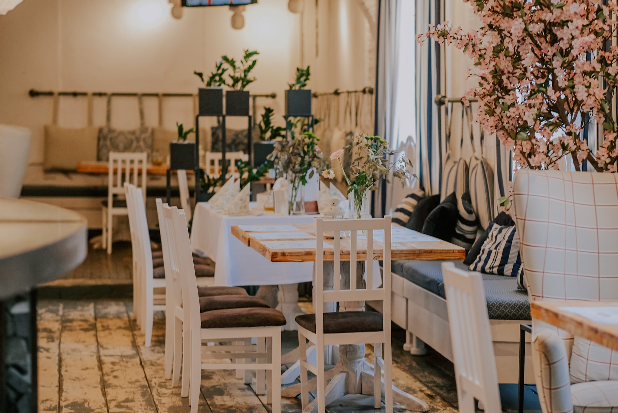 Uroczysta aranżacja wnętrza restauracji: białe krzesła, drewniane stoły z dekoracjami kwiatowymi i roślinnymi, tworzą przytulną atmosferę.