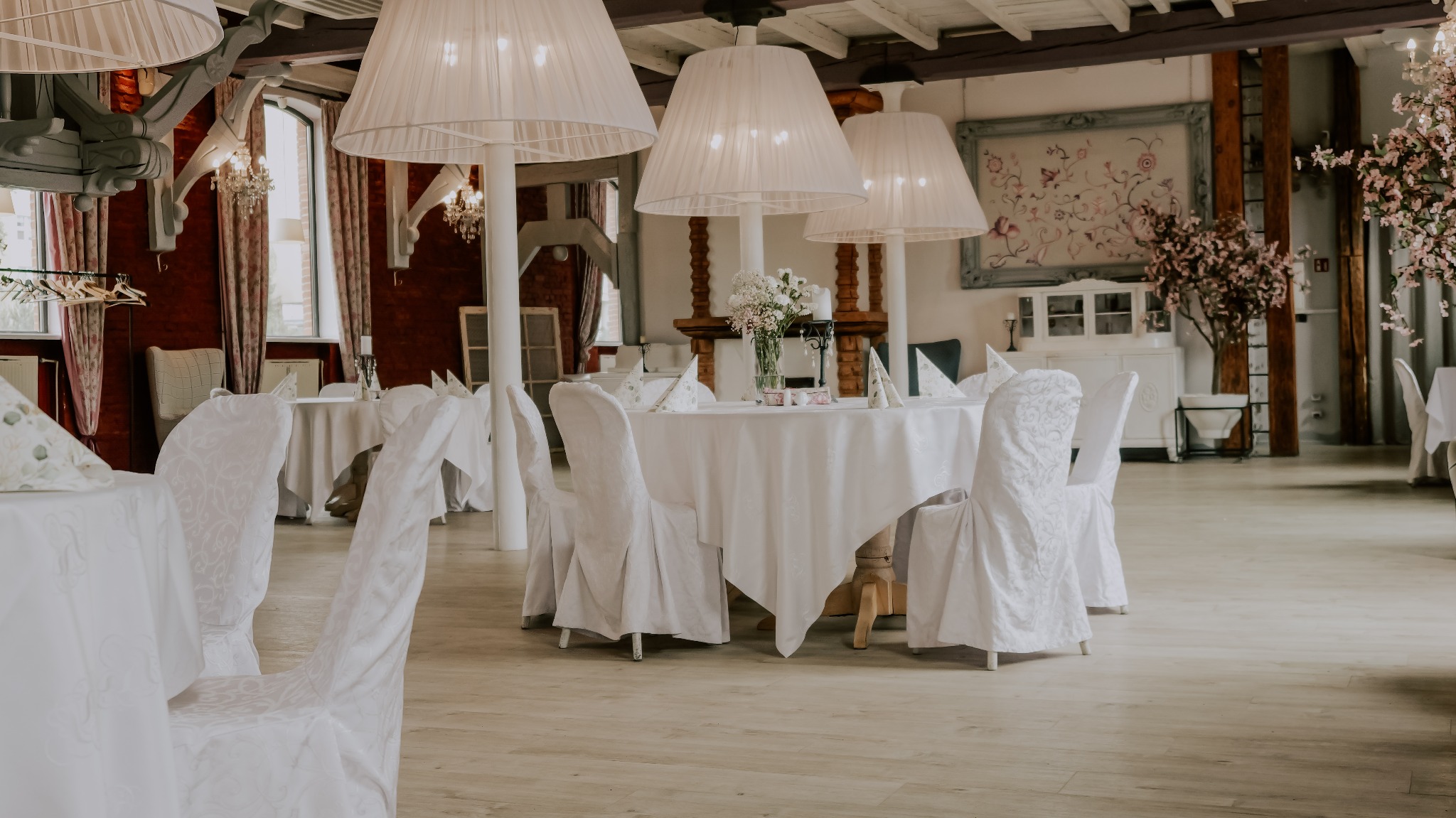 Elegancko nakryte stoły z białymi obrusami i dekoracjami kwiatowymi w przestronnej sali bankietowej z drewnianą podłogą, gotowej na przyjęcie.