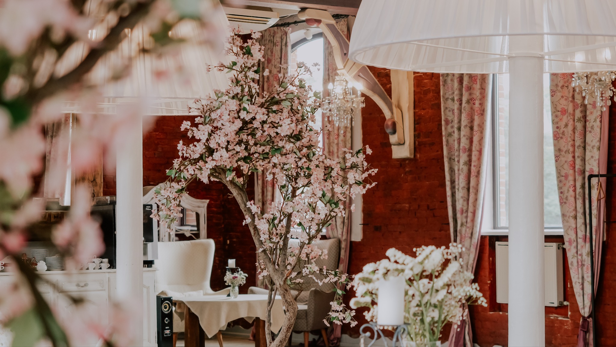 Dekoracja sali z kwitnącą wiśnią na stole i bukietami, ceglana ściana i żyrandol w tle, tworzą elegancką aranżację eventową.