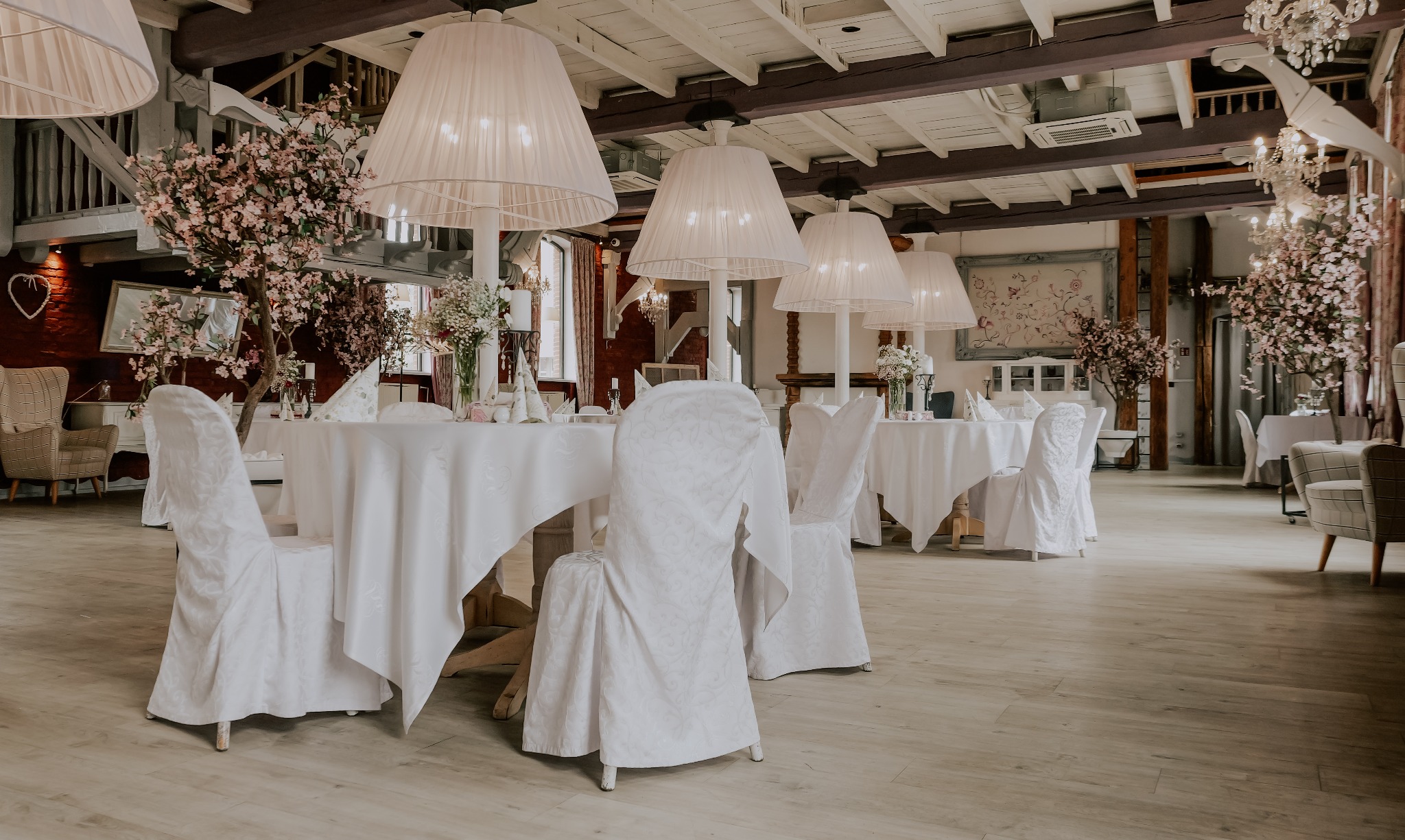 Elegancka sala bankietowa z białymi obrusami, krzesłami w pokrowcach i kwiatowymi dekoracjami. W tle widoczne dekoracyjne lampy i elementy rustykalnego wystroju.