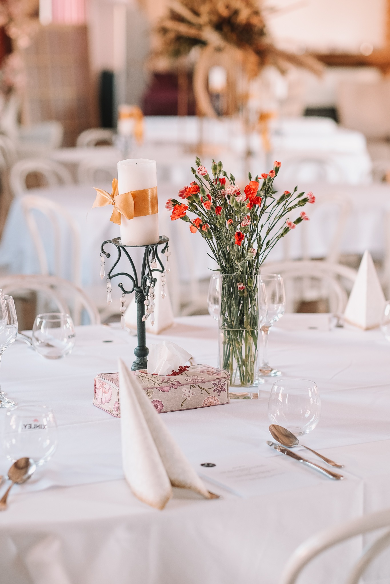 Dekoracja stołu: świeca ze wstążką, wazon z czerwonymi kwiatami, elegancka zastawa i serwetki na białym obrusie, w tle krzesła i elementy dekoracyjne sali.