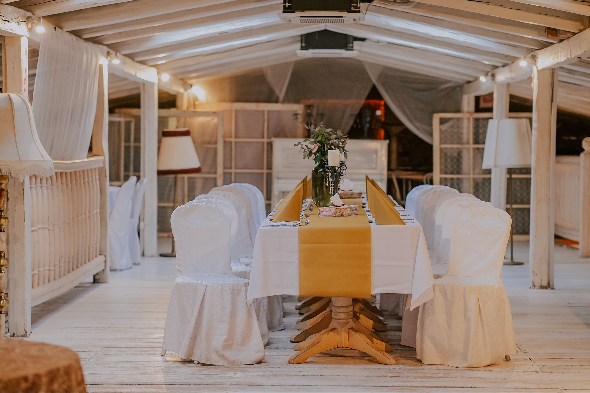 Uroczysty stół z białym obrusem i złotym bieżnikiem, nakryty dla gości, w jasnym wnętrzu z drewnianymi elementami i dekoracyjnymi lampami.