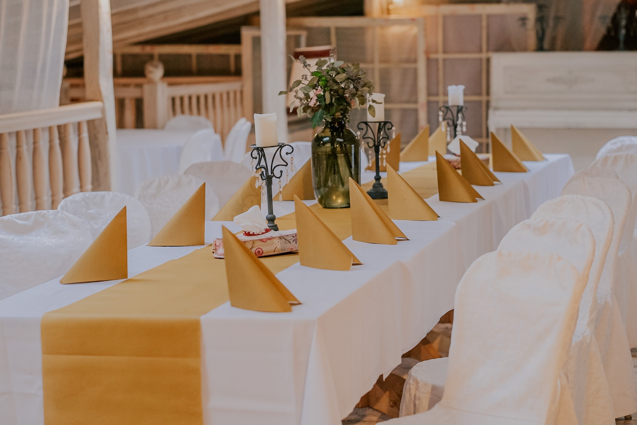 Uroczysty stół z białym obrusem, złotym bieżnikiem i serwetkami, dekorowany świecznikami i kwiatami w butelce, przygotowany na przyjęcie.