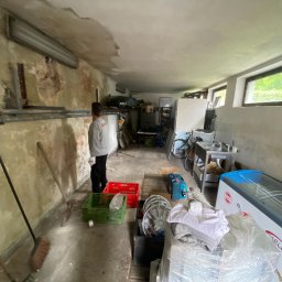 WORKMATES SP Z O O - Przygotowania do remontu: mężczyzna w szarej bluzie stoi w pomieszczeniu z odrapanymi ścianami, widoczne narzędzia, skrzynki i sprzęty.