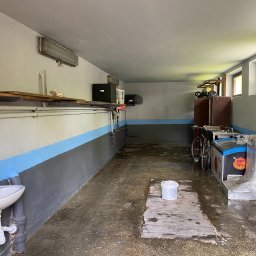 WORKMATES SP Z O O - Piwnica po malowaniu ścian na szaro-biało z niebieskim pasem. Widoczna umywalka, okna i sprzęty. Na podłodze stoi wiadro z farbą.