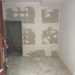 WORKMATES SP Z O O - Pokój w trakcie szpachlowania, widoczne ślady po łatach na ścianach i podkład na podłodze. Otwarta, ciemna stolarka drzwiowa z widocznym wiadrem i narzędziami.