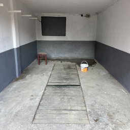 WORKMATES SP Z O O - Wnętrze garażu po malowaniu: ściany dwukolorowe (szaro-białe), drewniana podłoga z otworem, wiadro z farbą, krzesło. Świeże malowanie, surowy, minimalistyczny styl.