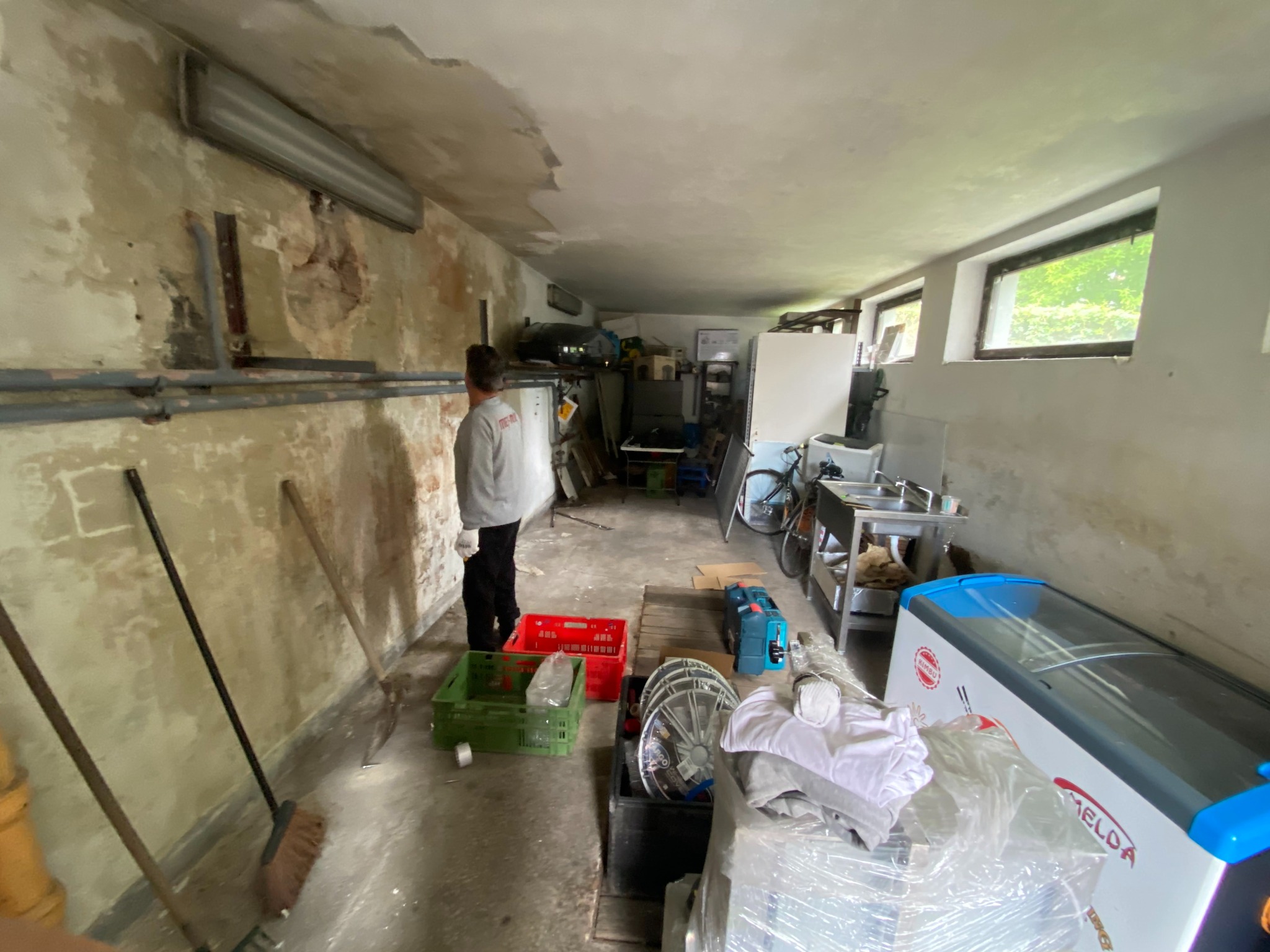 Przygotowania do remontu: mężczyzna w szarej bluzie stoi w pomieszczeniu z odrapanymi ścianami, widoczne narzędzia, skrzynki i sprzęty.